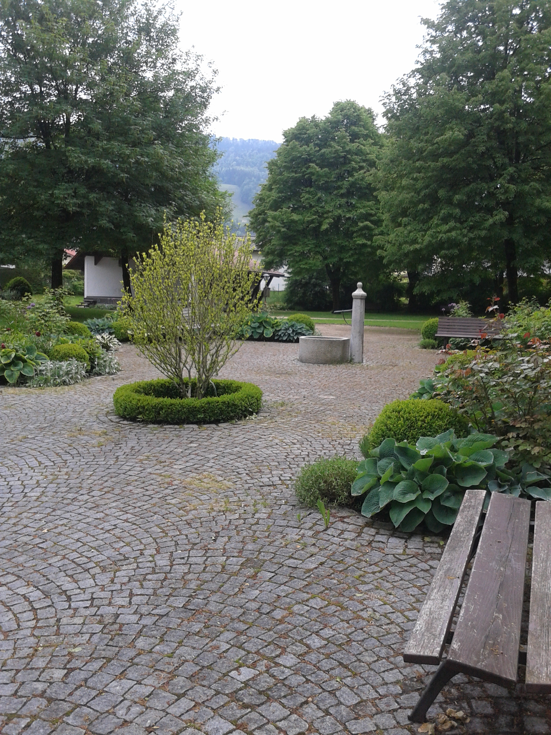 Zu sehen ist ein gepflasterter Platz, auf dem sich kleine grüne Inseln befinden. Sträucher und Beete sind darauf angelegt. Ringsherum stehen Holzbänke. Am Ende des Platzes sieht man einen kleinen, steinernen runden Brunnen mit Wasserhahn. Im Hintergrund liegen Grünflächen mit großen Bäumen, hinter diesen versteckt sich ein Haus.
